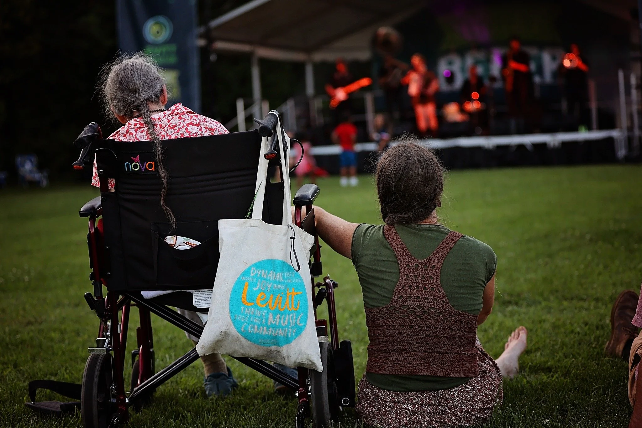 An elderly woman in a wheelchair and a young woman sitting on the grass watching a concert on an outdoor stage at dusk.