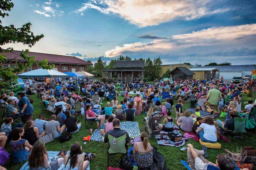 Outdoor community gathering with people seated on blankets and lawn chairs facing a small stage, under a partly cloudy sky with buildings and trees in the background.