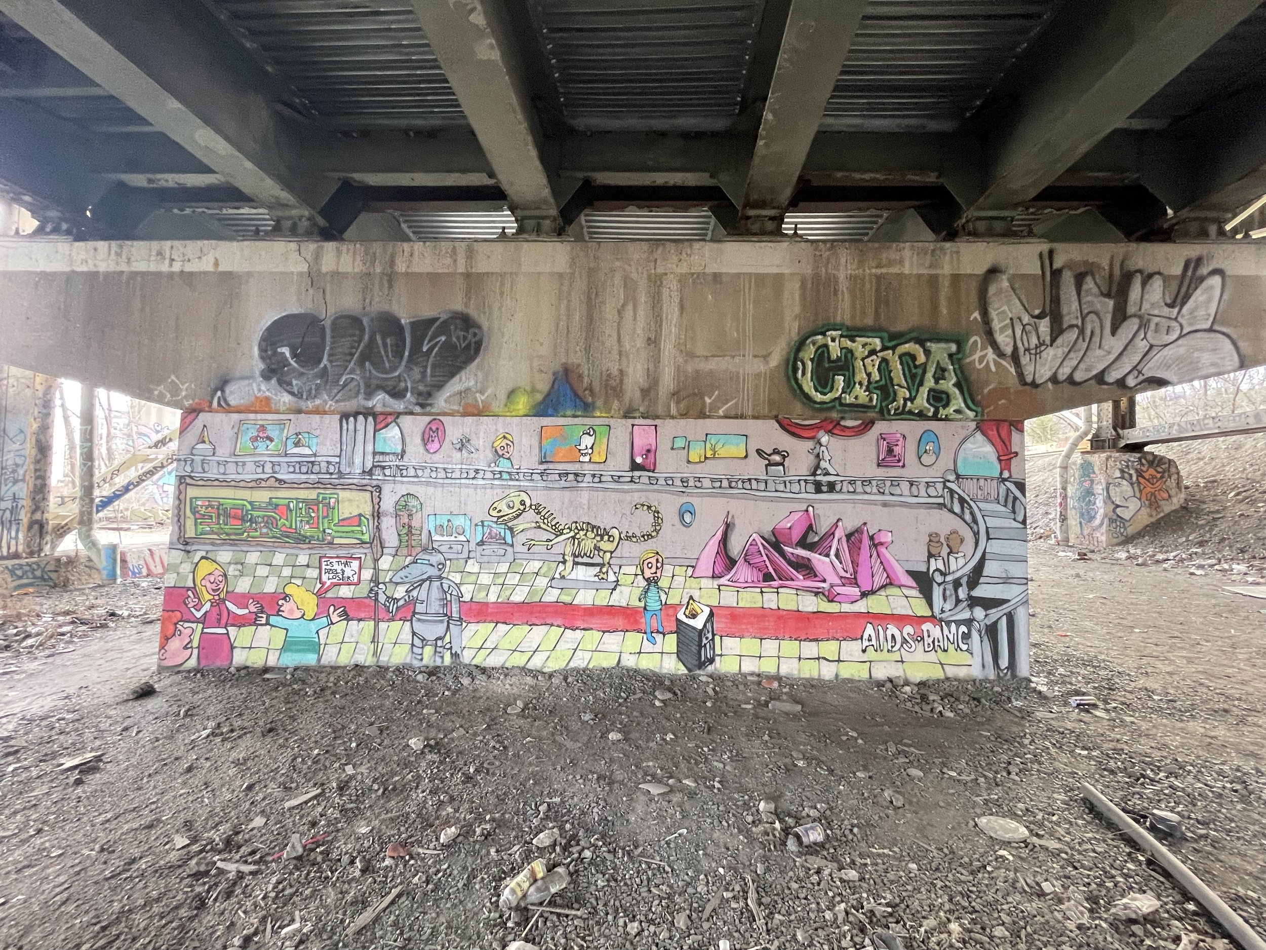 Colorful graffiti mural under a bridge depicting cartoon characters, a dinosaur skeleton, and various abstract designs.