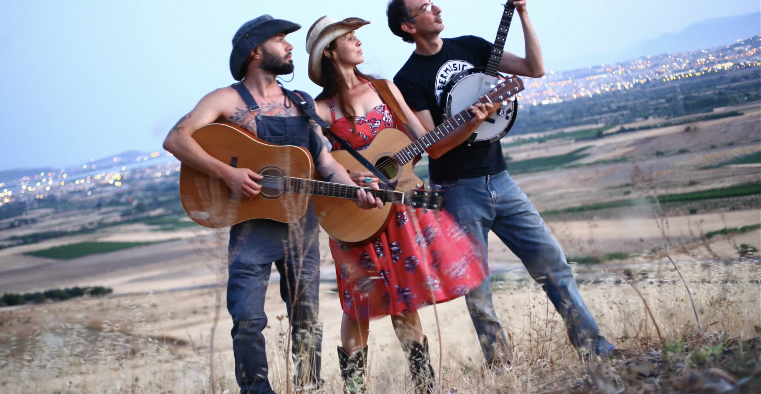 Three people playing guitars outdoors on a hill with a cityscape in the background during evening.