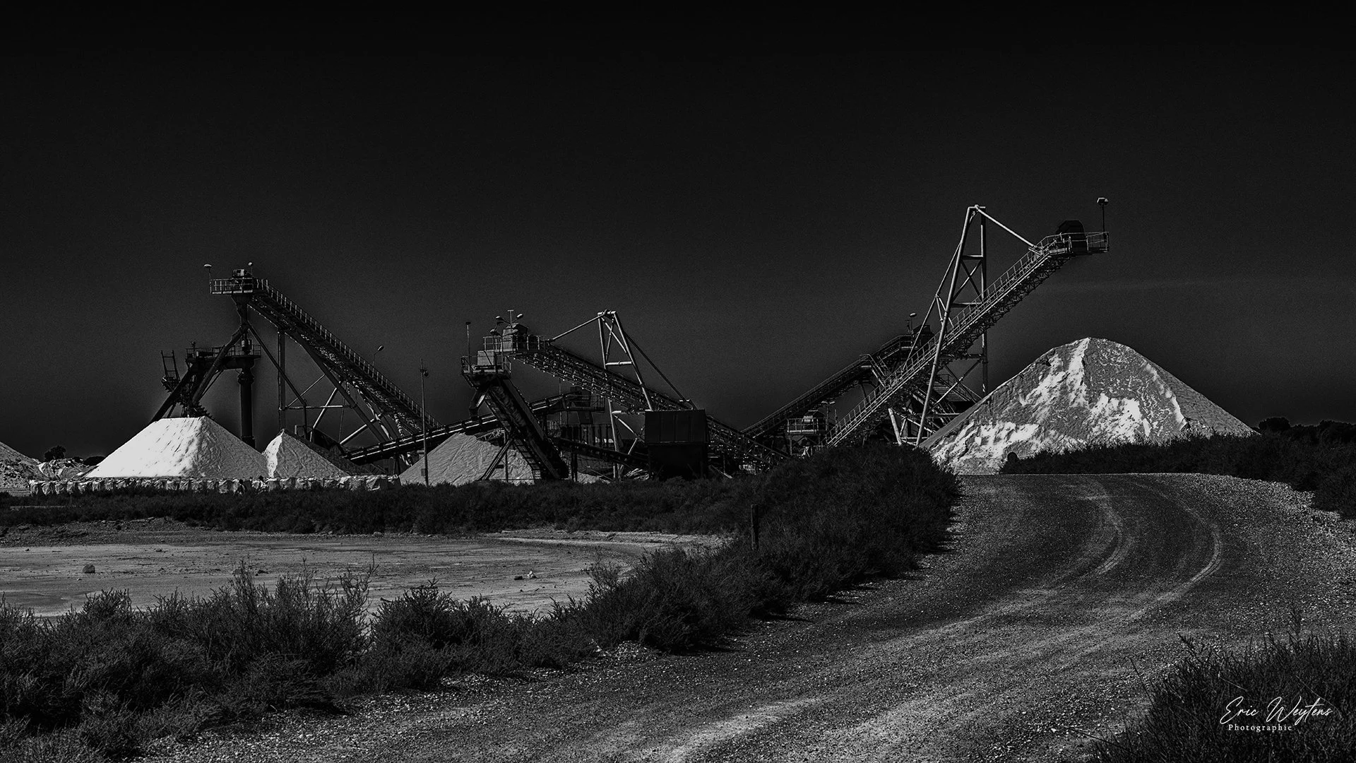 Photographie en noir et blanc d'une carrière avec des tas de sel ou de pierre, des équipements d'exploitation, et une montagne ou un mont enneigé en arrière-plan, sous un ciel sombre.