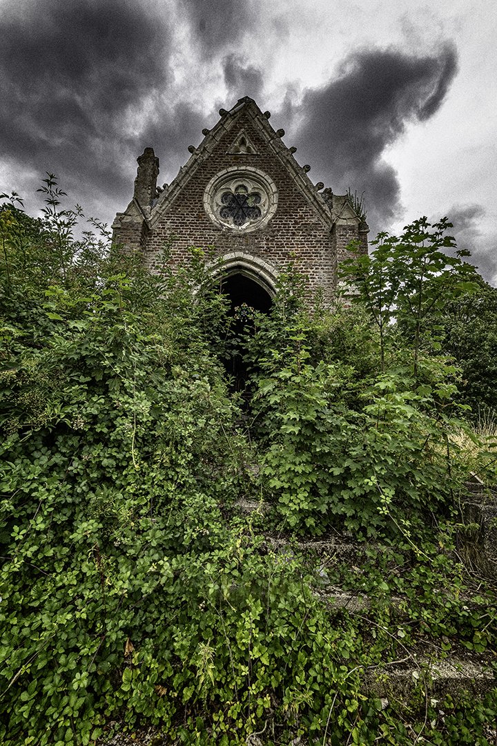 Une vieille église en ruine, envahie par la végétation, avec un ciel nuageux en arrière-plan.