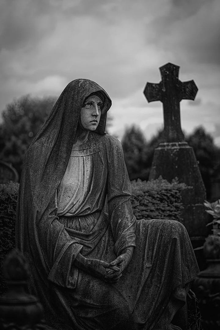Statue en pierre d'une femme assise dans un cimetière, avec une croix en pierre en arrière-plan, sous un ciel nuageux.