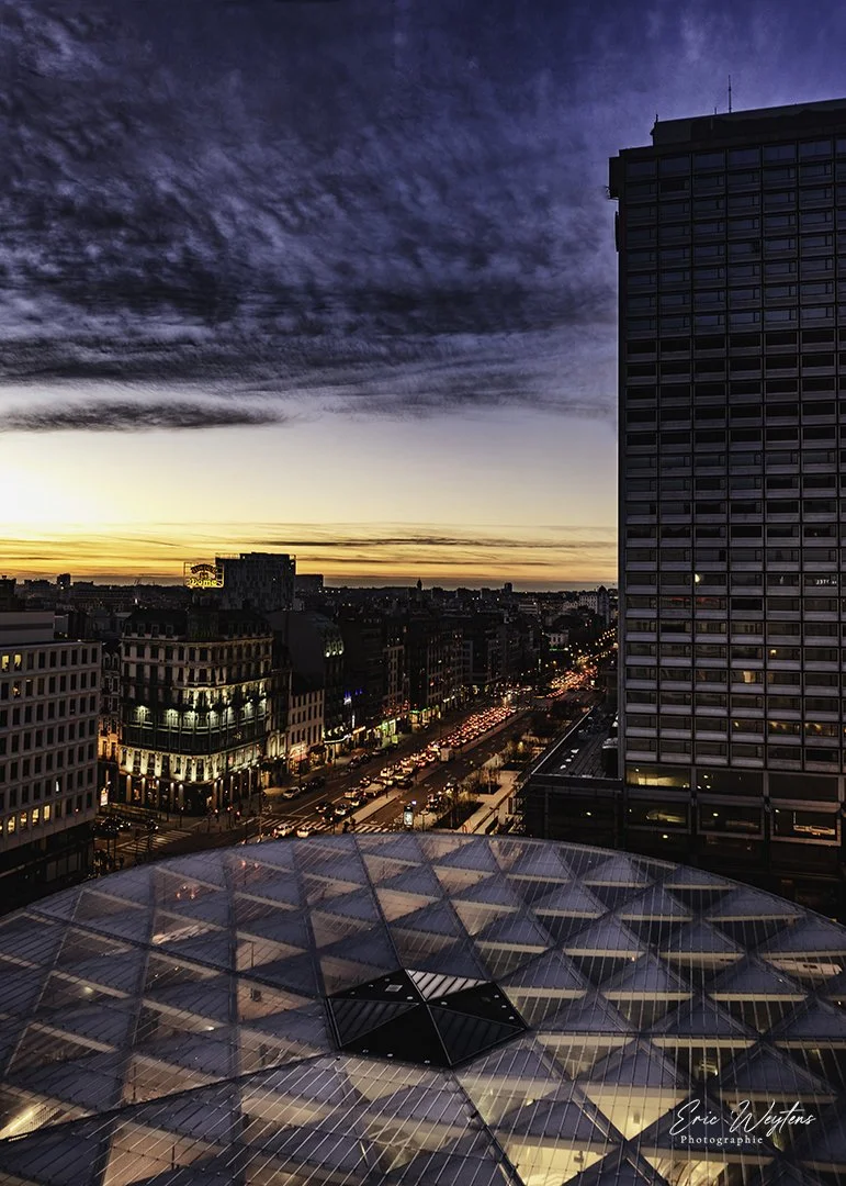 Vue d'une ville la soirée avec un ciel coloré au coucher du soleil, un grand bâtiment moderne avec de nombreux fenêtres et un toit en forme géométrique en verre