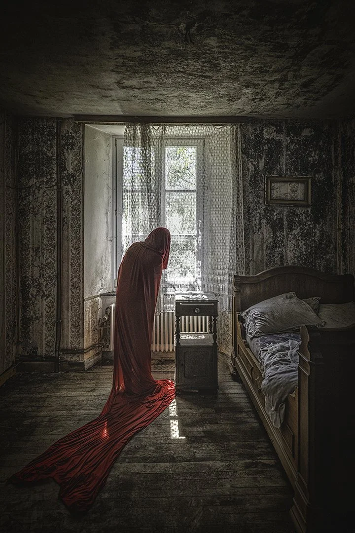 Une pièce abandonnée avec des murs vieux et dégradés, une fenêtre laissant passer la lumière, une femme en robe rouge long et fluide se tenant devant la fenêtre, avec le nu de la robe étalé sur le sol.