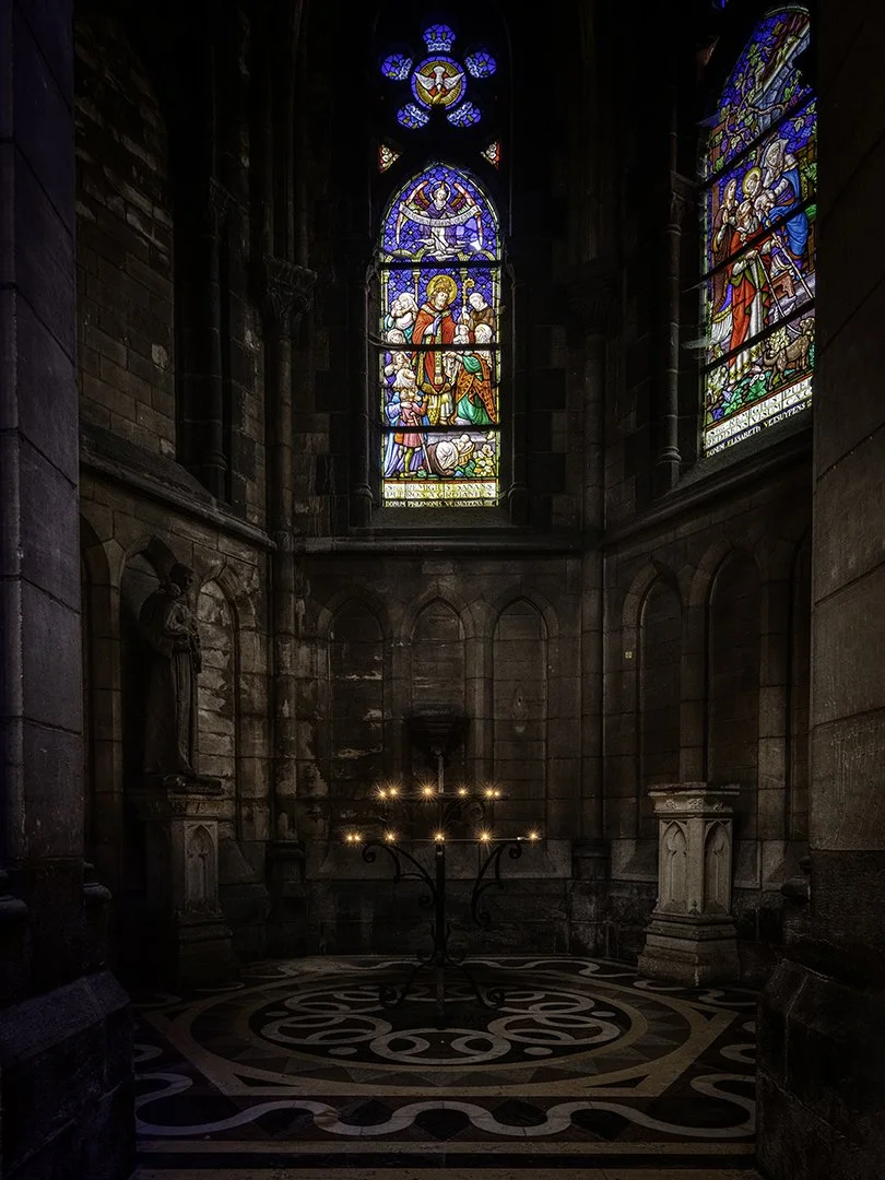Intérieur d'une église avec des vitraux colorés représentant des scènes religieuses, une statue de saint, et un chandelier à bougies au centre sur un sol ornée de motifs.