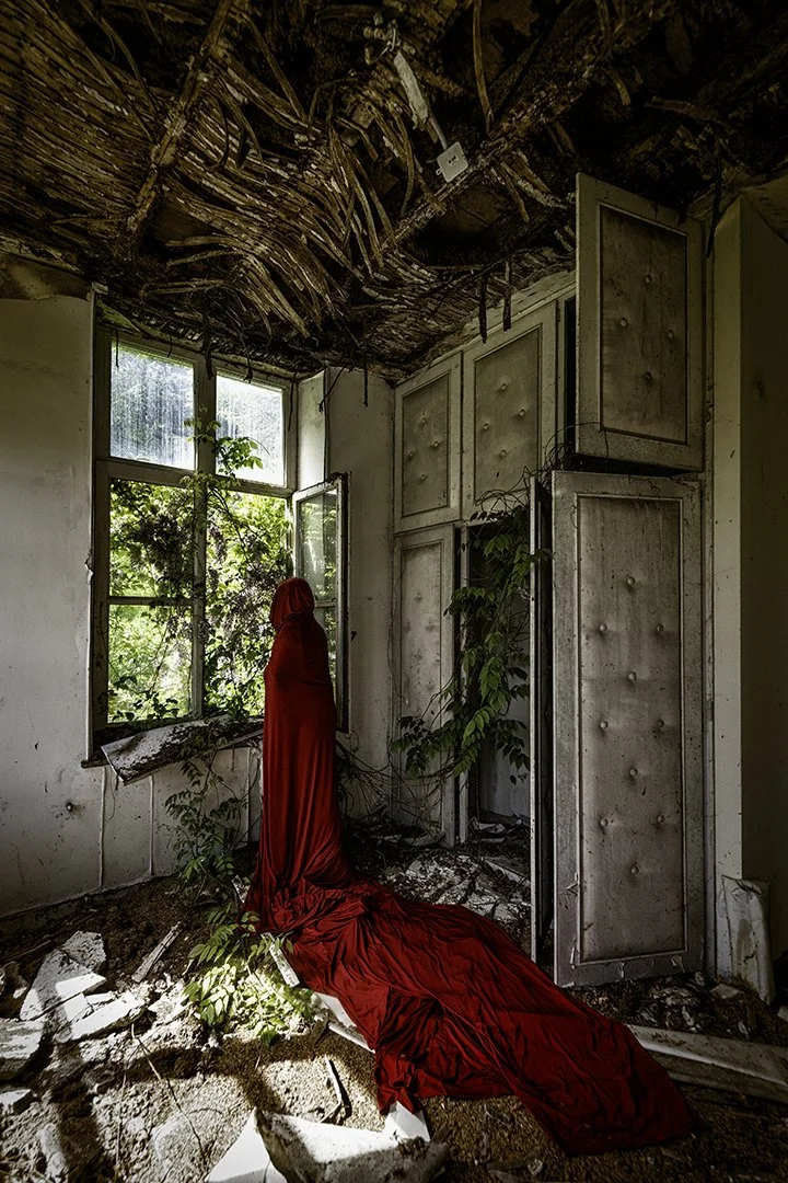 Une femme en robe rouge dans une pièce abandonnée avec un plafond endommagé, des fenêtres cassées laissant entrer la lumière, des plantes qui poussent à l'intérieur, et du débris au sol.