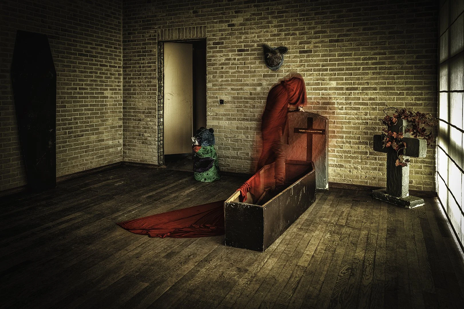 Un espace intérieur avec murs en briques et sol en bois, décoré avec une croix en bois sur un meuble noir, une sculpture de pierres avec des fleurs dans un pot en pierre, et une personne vêtue de rouge dont le corps semble flou. Il y a aussi une scul