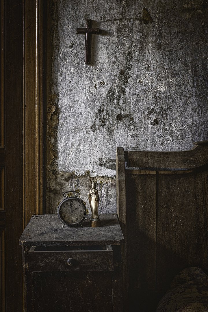 Une vieille chambre ou un espace intérieur avec un mur en pierre et une croix en bois, une table ou un meuble avec une horloge ancienne, une petite statue dorée, et un lit partiellement visible sur le côté droit.