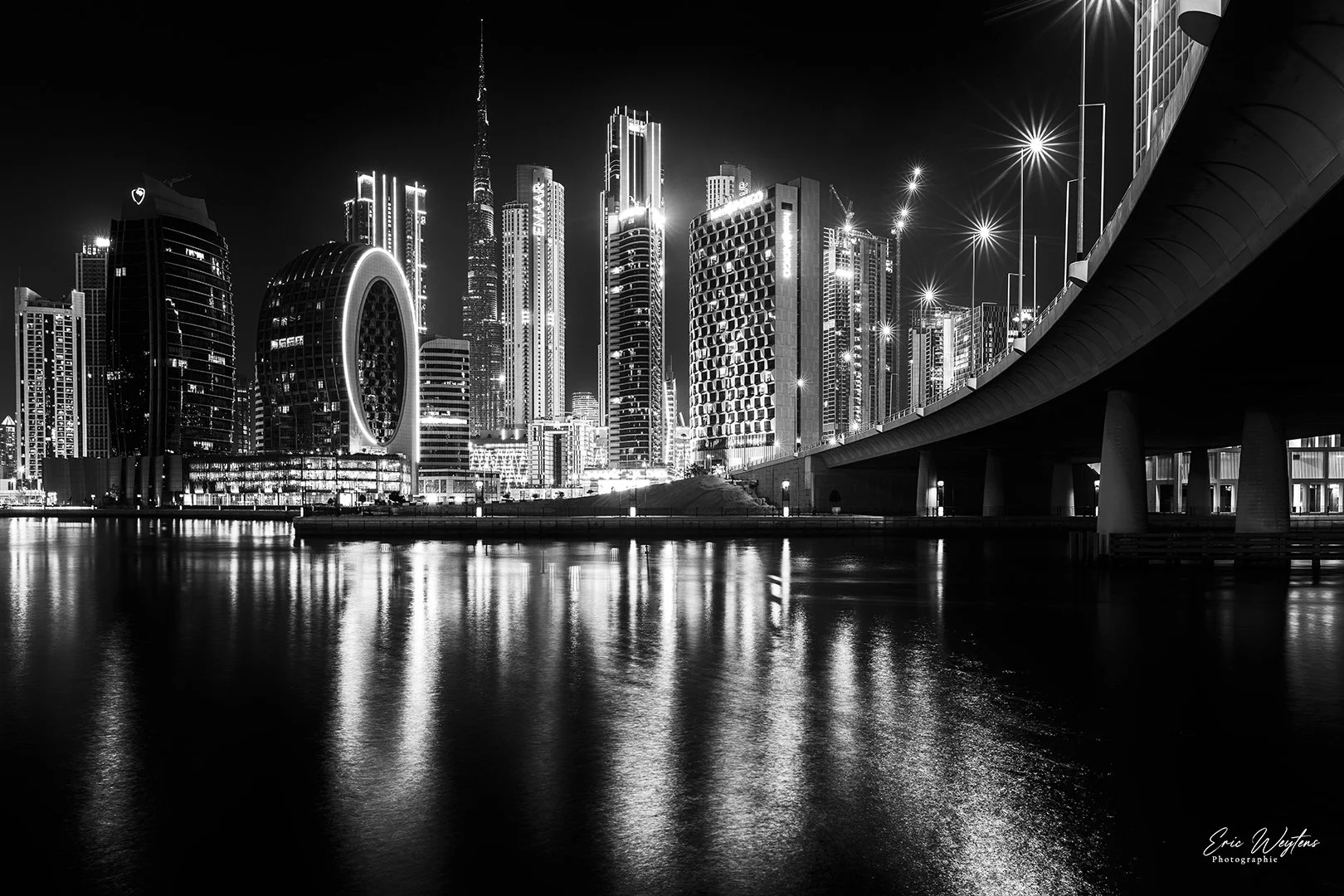 Une ligne d'horizon de gratte-ciel modernes illuminés la nuit, reflétés dans l'eau calme au premier plan, avec un pont suspendu. En bas à droite, le nom du photographe, Eric Weigand.