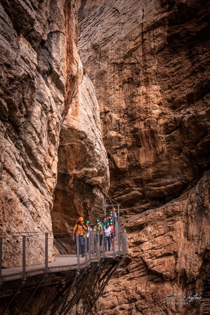 Groupe de personnes en randonnée dans une gorge rocheuse, portant des casques de sécurité et marchant sur un pont suspendu en métal.