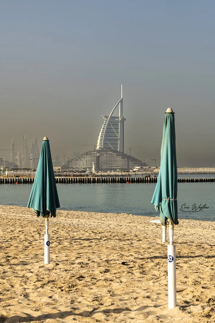 Plage avec deux parasols turquoise et la skyline de Dubaï en arrière-plan, dont le célèbre hotel Burj Al Arab.