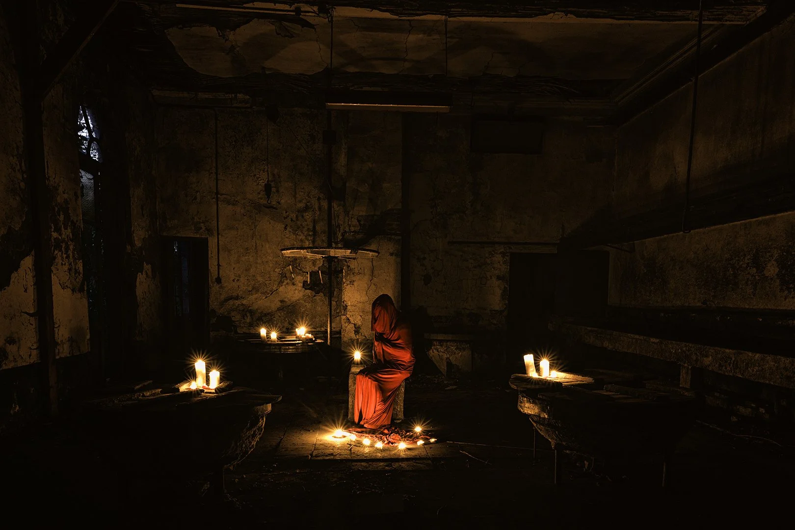Une pièce sombre et ancienne avec des murs en pierre, éclairée par plusieurs bougies. Au centre, une personne vêtue d'une robe rouge est assise, entourée de bougies allumées sur le sol et des tables en pierre. L'ambiance est mystérieuse et sombre.