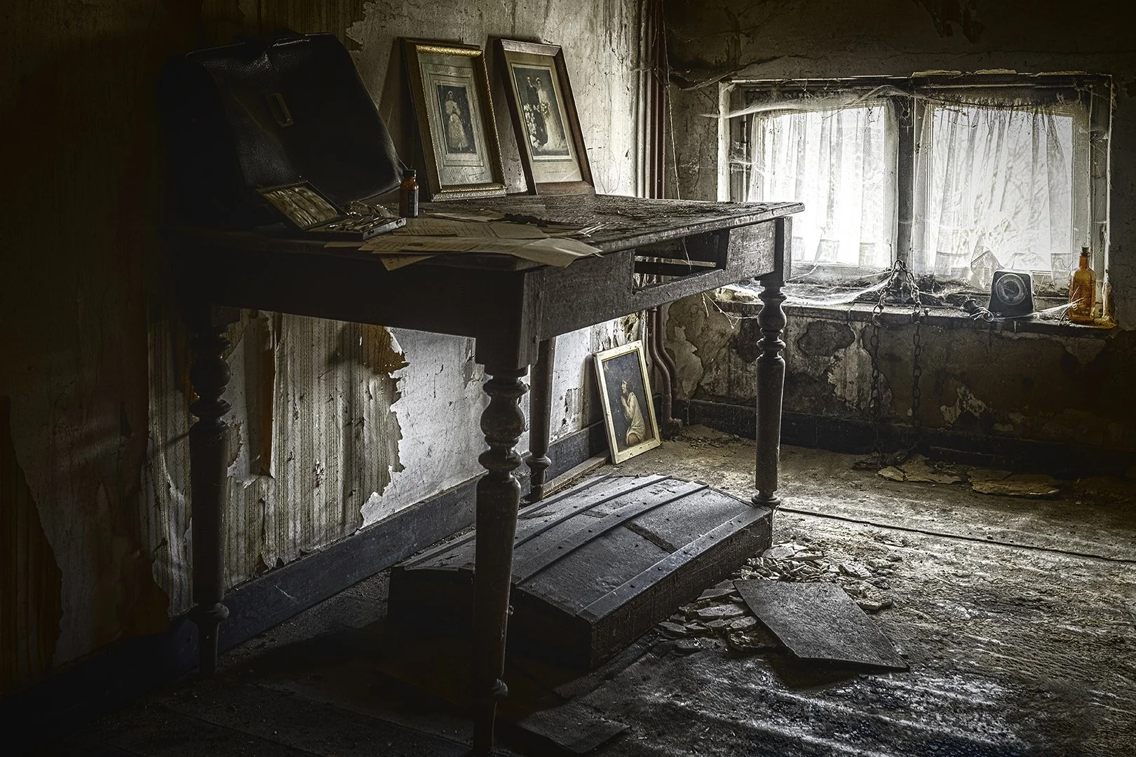 Intérieur d'une pièce abandonnée avec un bureau en bois usé, des cadres photo anciens, des papiers, une fenêtre sale avec des rideaux en toile d'araignée, et un sol en désordre avec des débris et un coffre en bois.