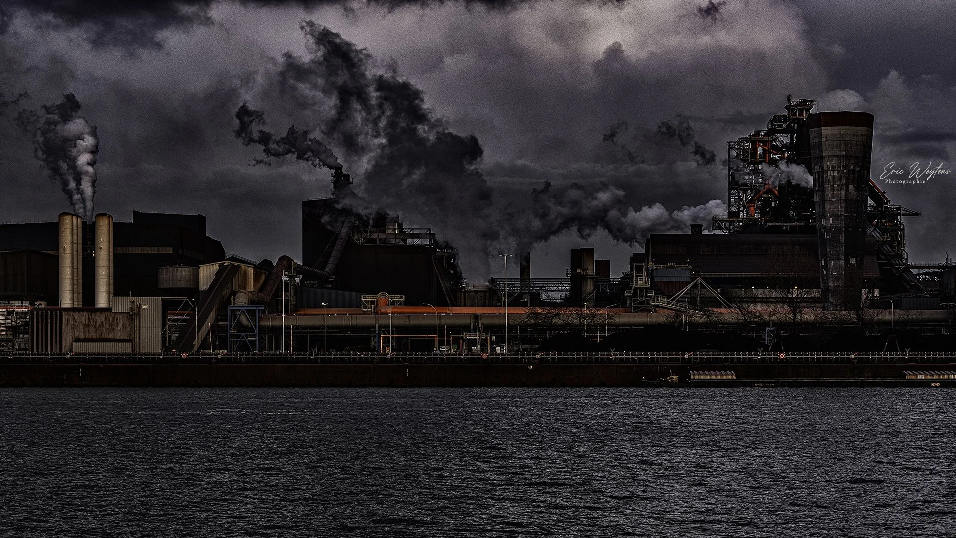 Une centrale électrique industrielle produisant beaucoup de fumée noire, avec un corps d'eau devant et un ciel nuageux.