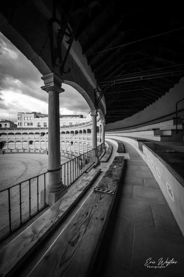 Une vue d'un colisée en noir et blanc avec des arches, colonnes et bancs en bois sur un côté, sous un ciel nuageux.