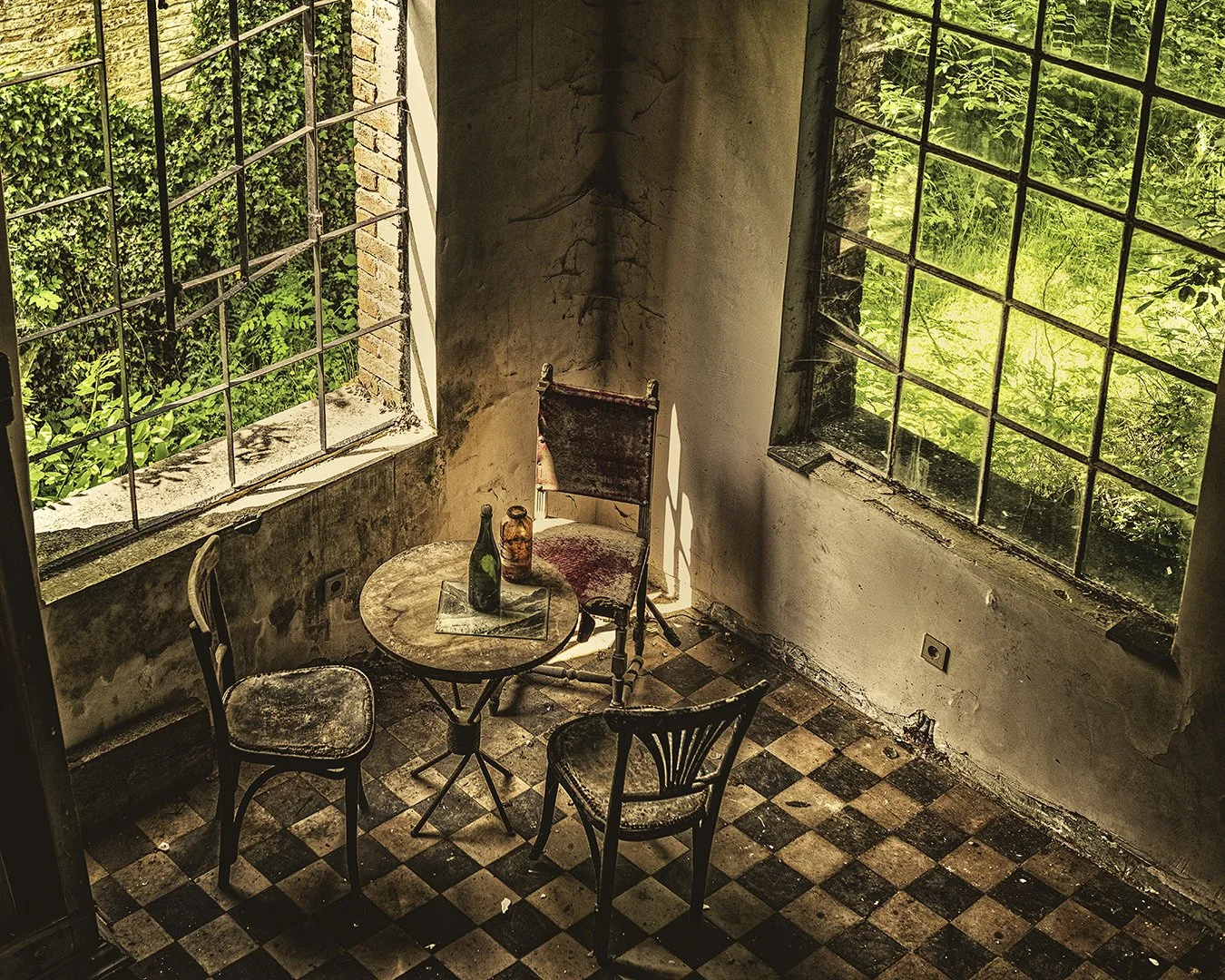 Une pièce abandonnée avec des murs en mauvais état, deux grandes fenêtres cassées laissant entrer la lumière et la verdure extérieure, un petit mobilier ancien avec une table en bois et trois chaises usées, un verre vert et un vase en verre sur la ta