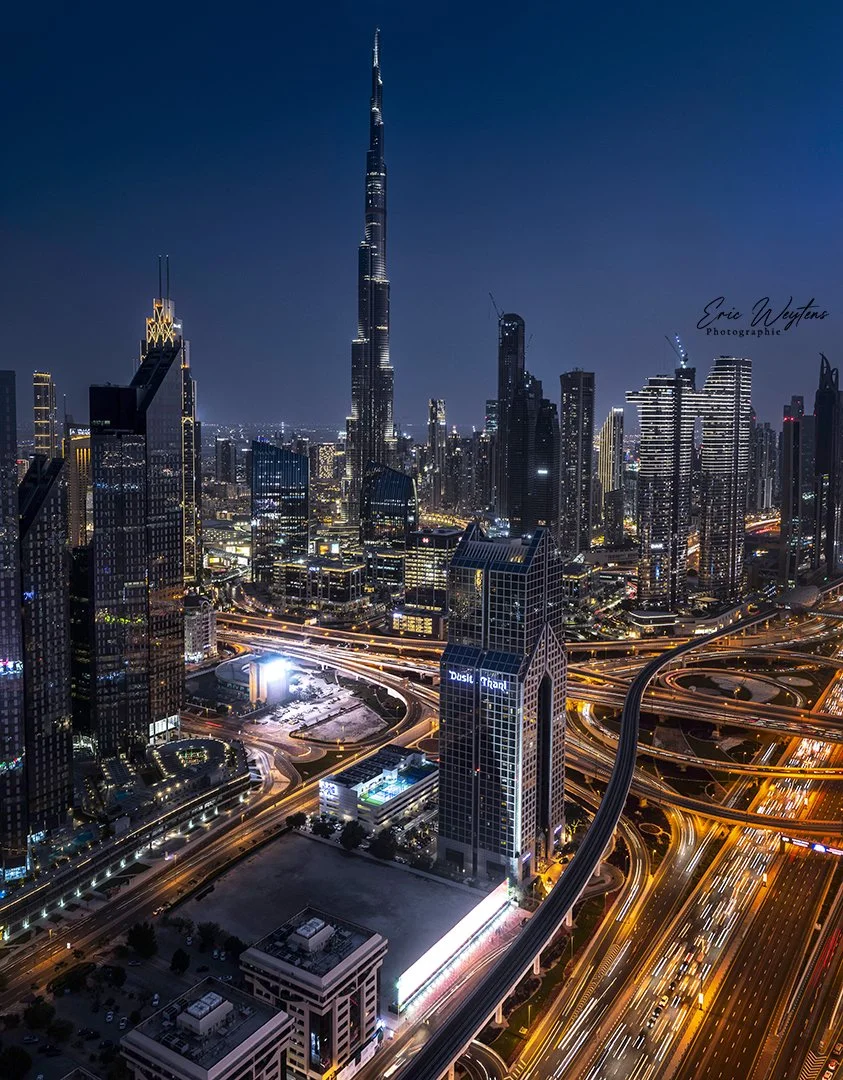Vue nocturne du skyline de Dubai avec la tour Burj Khalifa au centre, entourée de gratte-ciel illuminés et de routes éclairées entre les bâtiments.