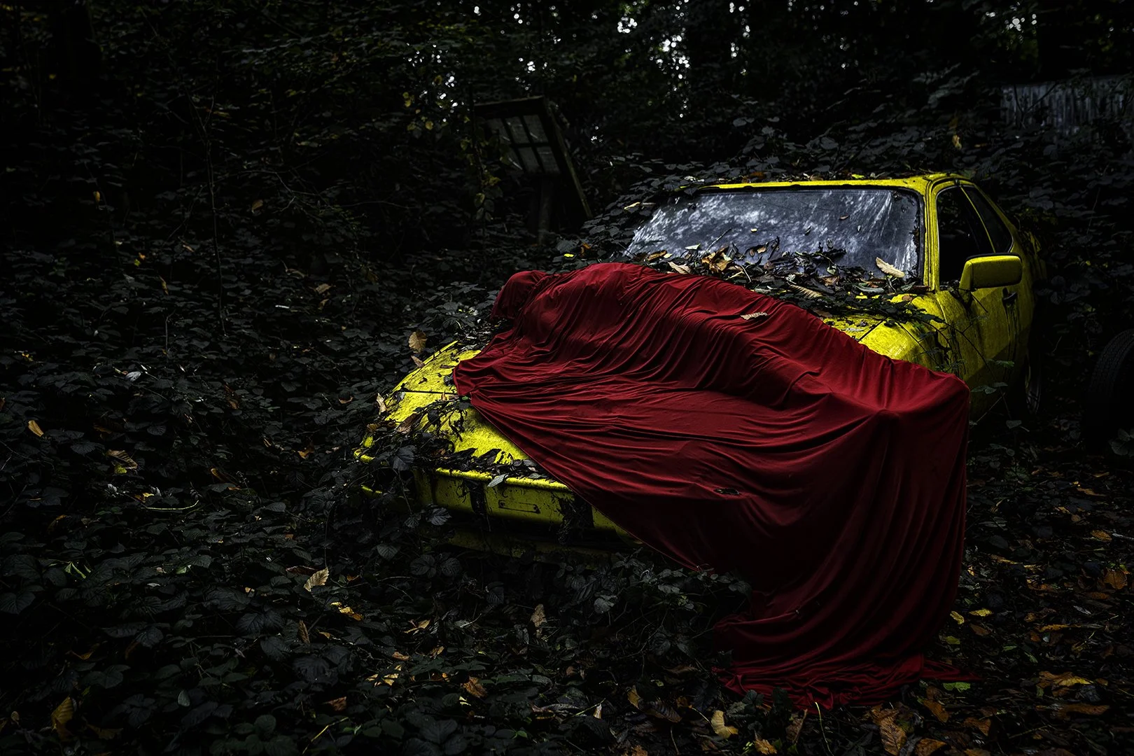Voiture abandonnée jaune dans une forêt, recouverte de feuilles et avec un drapé rouge dessus.