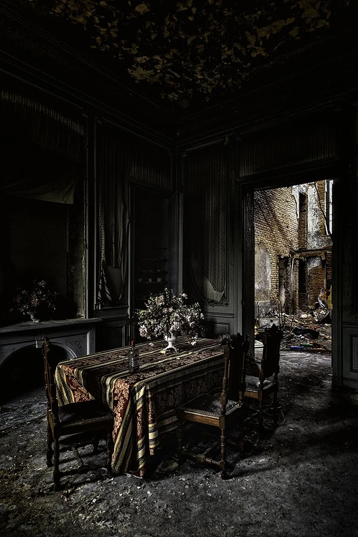 Une salle à manger abîmée avec un tableau couvert de poussière, un lustre cassé au plafond et des murs endommagés, vue d'une fenêtre ouverte sur une façade de bâtiment détruit.