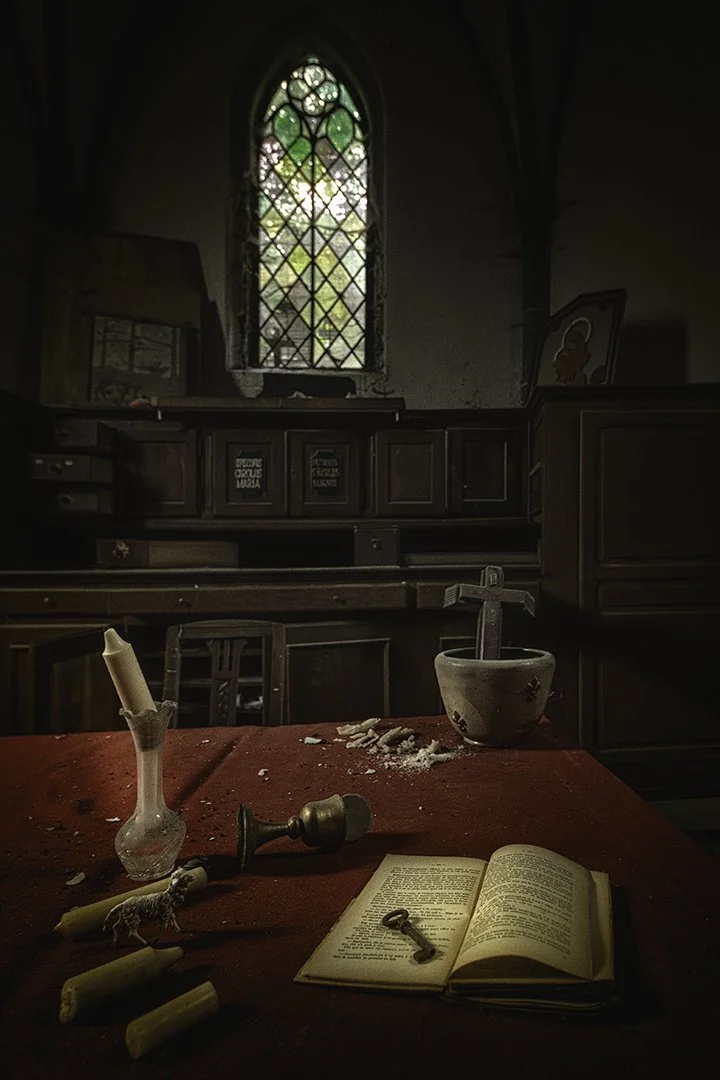 Une pièce sombre avec un vitrail coloré, un mobilier ancien, une table rouge couverte de vieux objets dont un livre ouvert, un chandelier, un verre avec une chandelle cassée, une sorte de crucifix, et une petite statue de mouton.