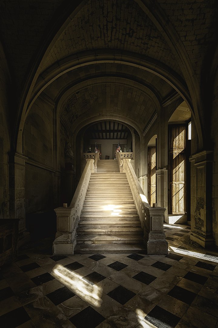 Escalier en pierre dans un hall ancien avec des arches et fenêtres en bois, illuminé par la lumière naturelle.