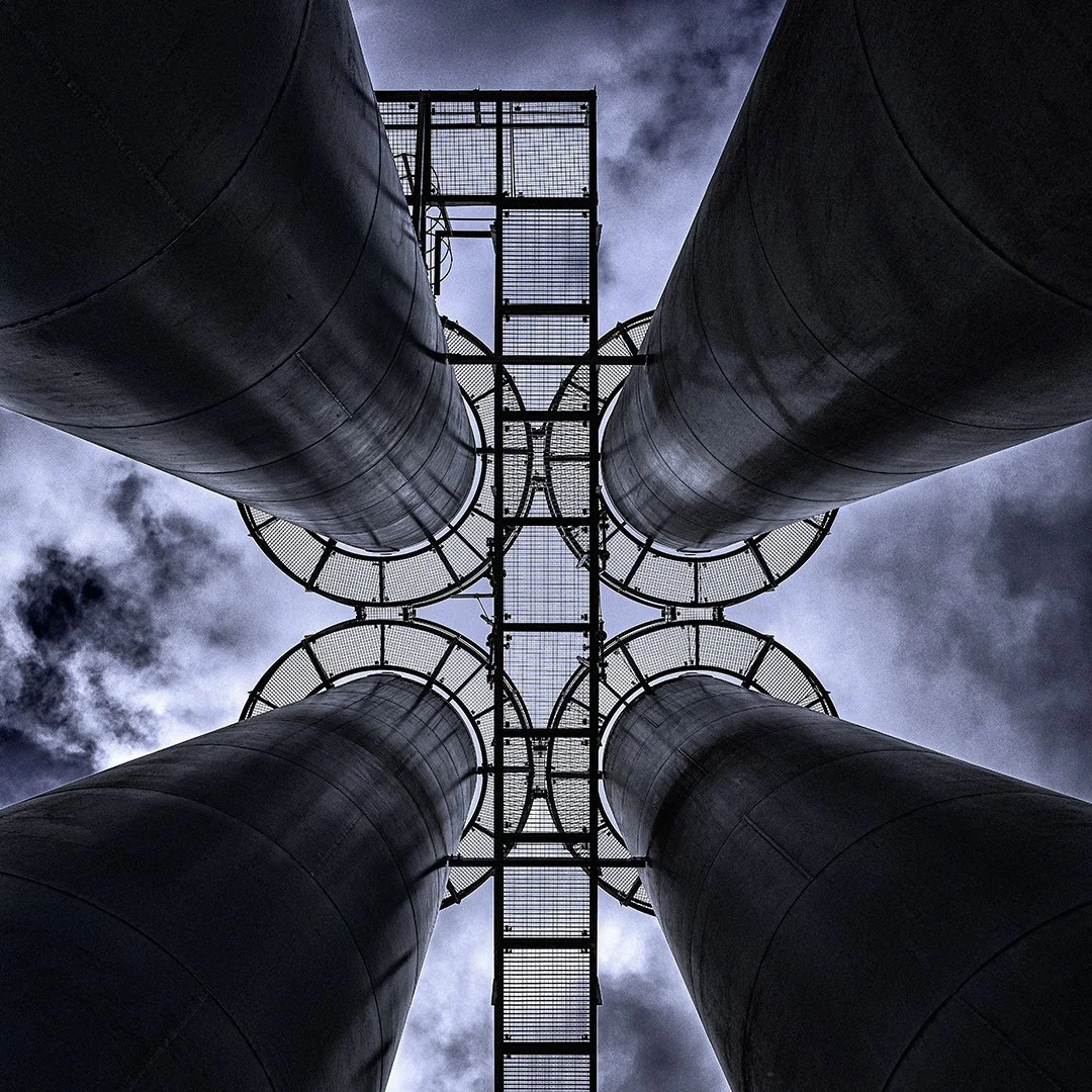 Vue ascendante de quatre grandes canalisations métalliques reliées par une plateforme métallique en haut, contre un ciel nuageux.