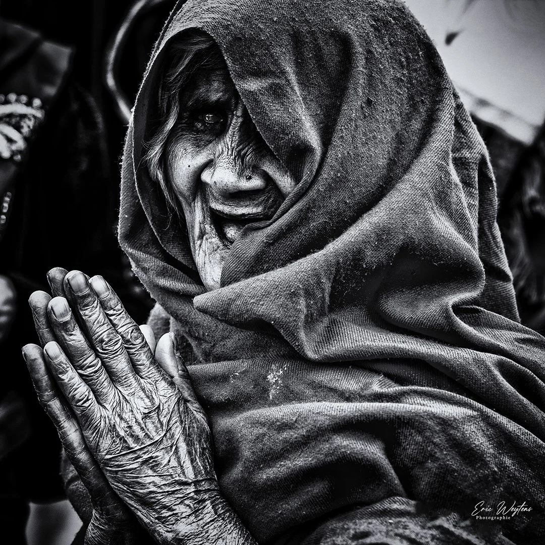 Une femme âgée avec un visage marqué et une peau ridée, portant un foulard sur la tête, fait un geste de prière avec ses mains jointes.