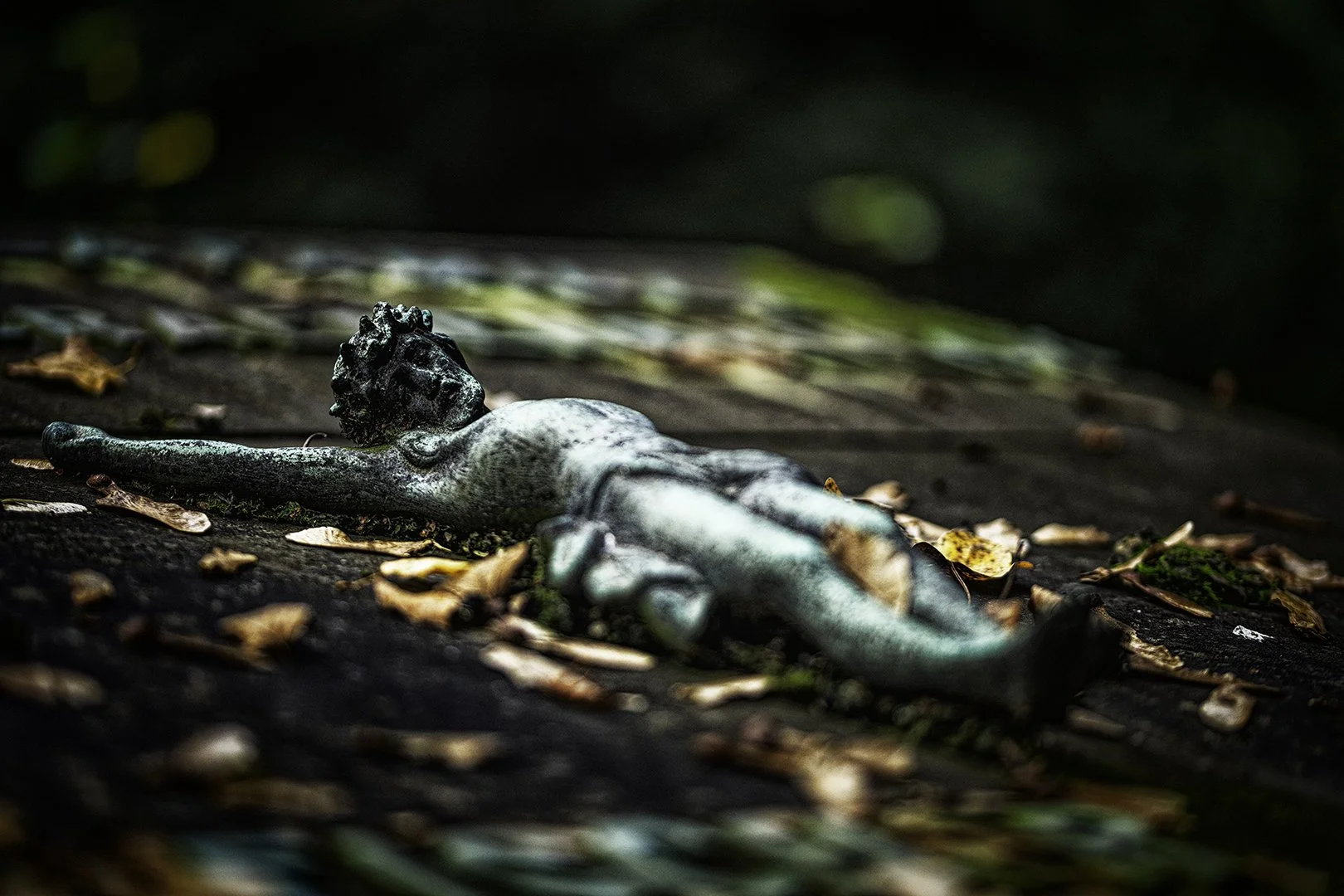 Une sculpture en pierre d'une figure humaine allongée sur le sol recouvert de feuilles mortes dans un environnement forestier sombre.