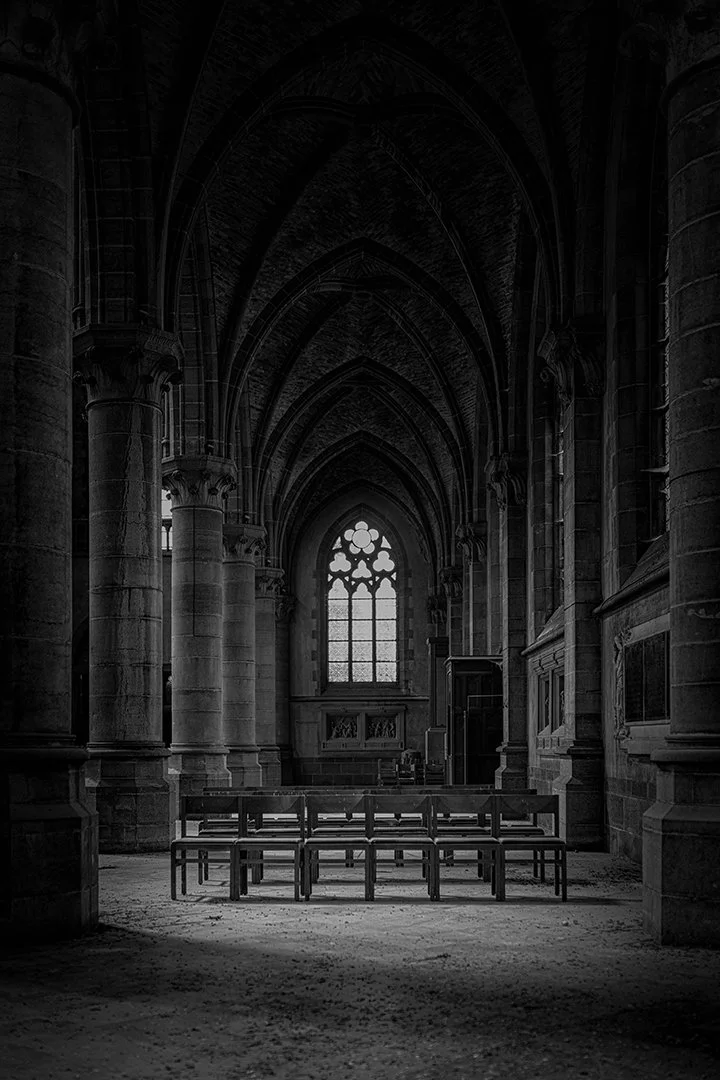 Intérieur d'une église gothique en noir et blanc avec des arches, des colonnes et un vitrail au fond, avec des chaises alignées au centre.