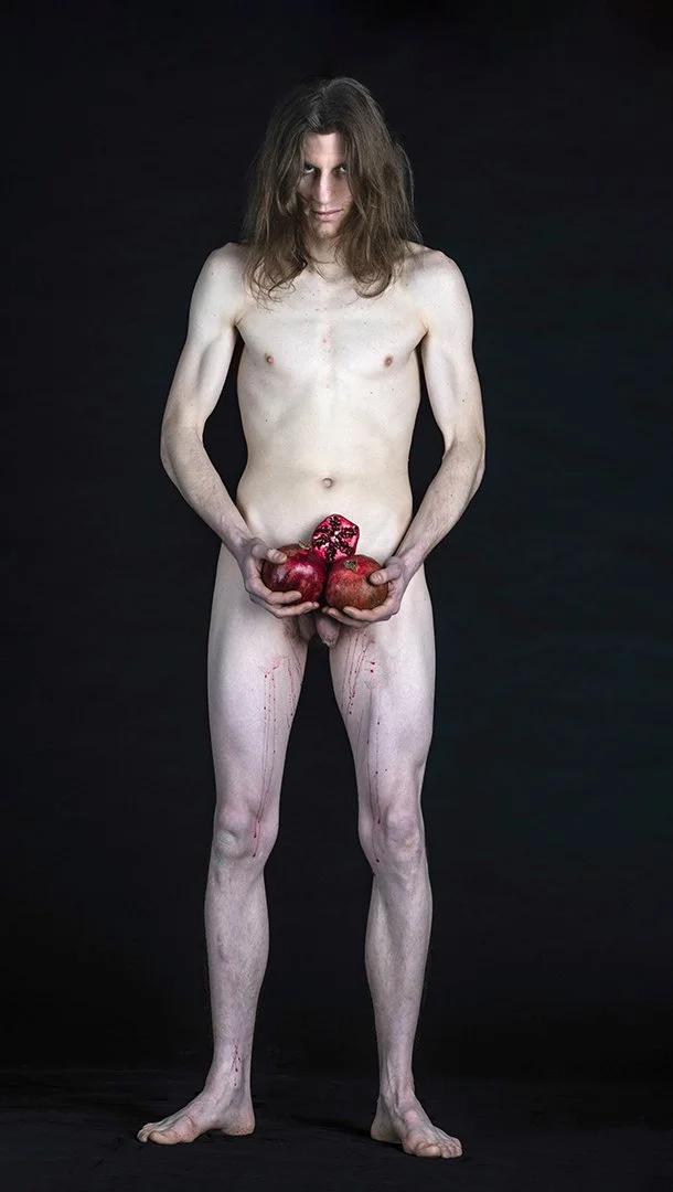 Un homme nu, yeux fixes, tenant trois grenades rouges, une de face et deux sur le côté, avec des éclaboussures de liquide rouge sur le corps, posé sur un fond noir.