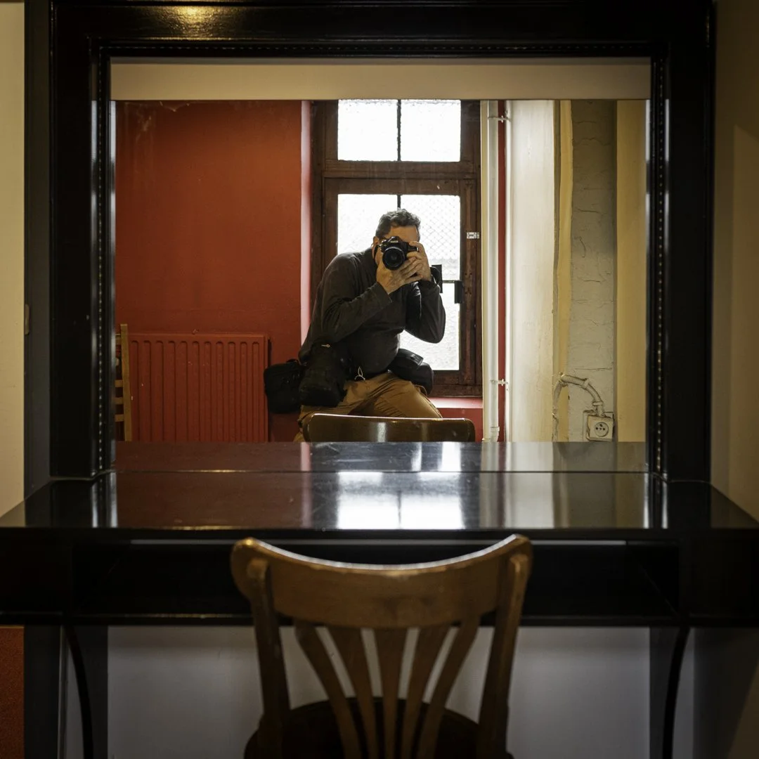 Un homme prenant une photo de lui-même dans un miroir, assis devant un bureau en bois au fond d'une pièce avec murs rouge et beige, derrière une fenêtre avec une grille.