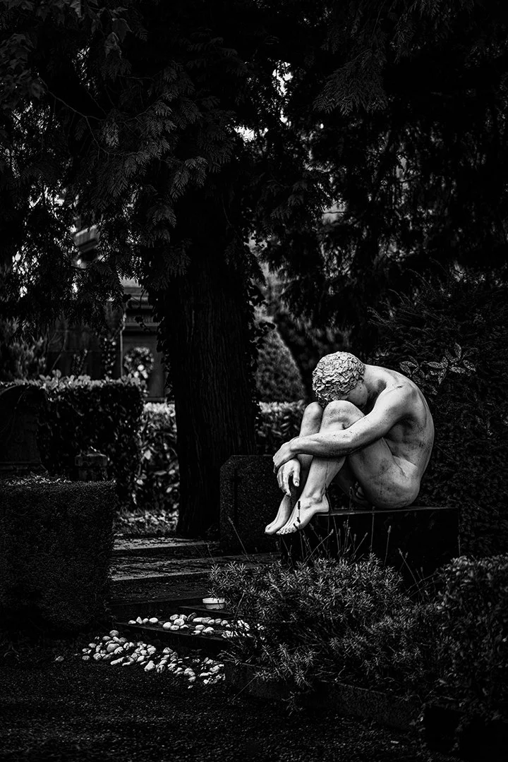 Statue en marbre d'une femme assise, le visage penché sur ses jambes dans un jardin entouré d'arbres et de buissons.