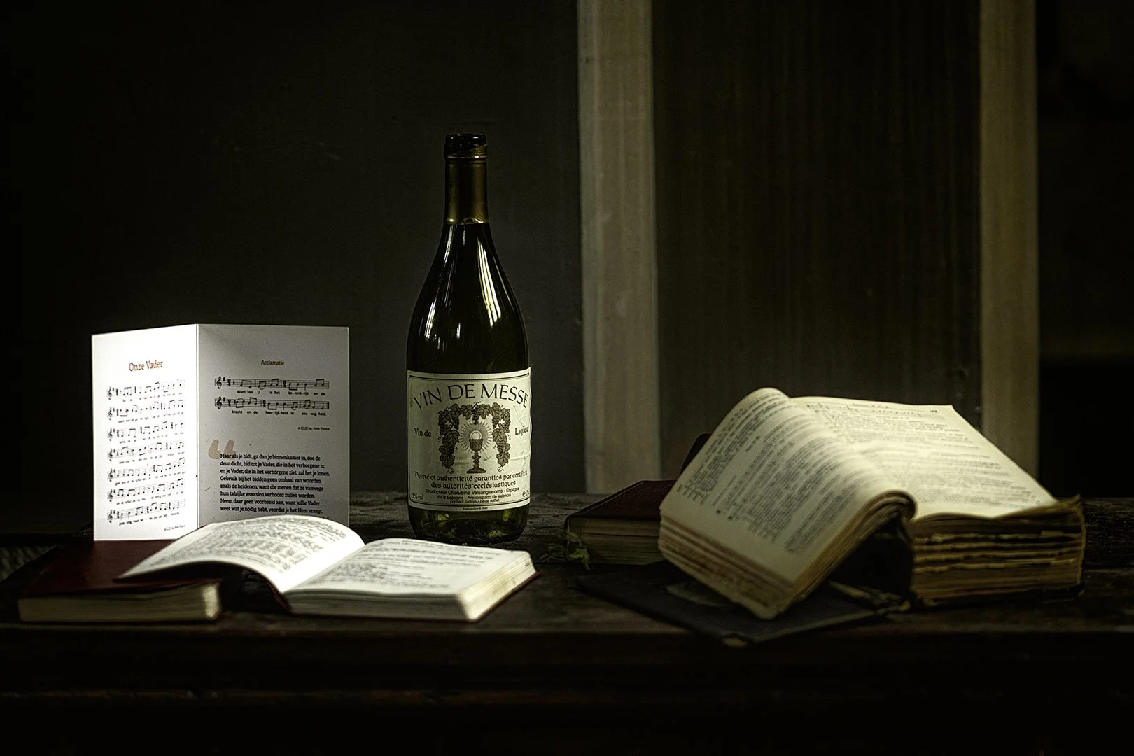 Étude religieuse avec livres anciens ouverts, une bouteille de vin, une brochure de musique, et une photographie religieuse sur une table en bois.
