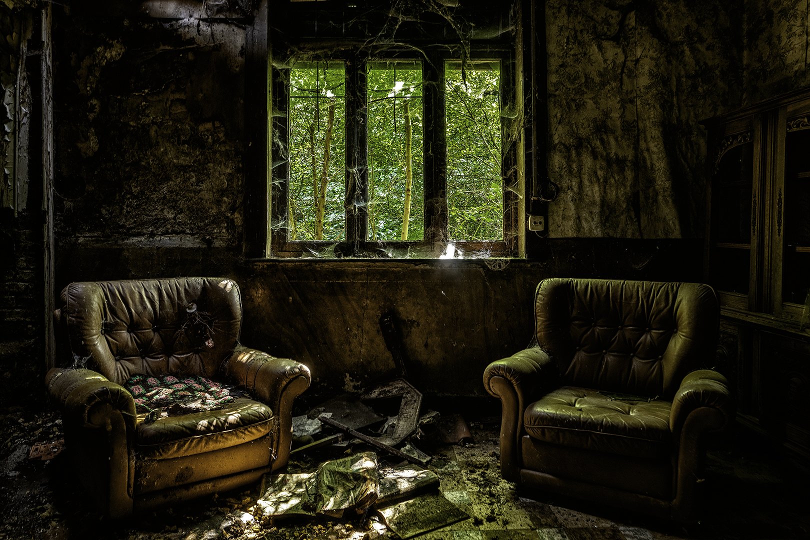 Intérieur d'une maison abandonnée avec deux fauteuils en cuir usés, une fenêtre laissant passer la lumière, des toiles d'araignées et des murs décrépis.