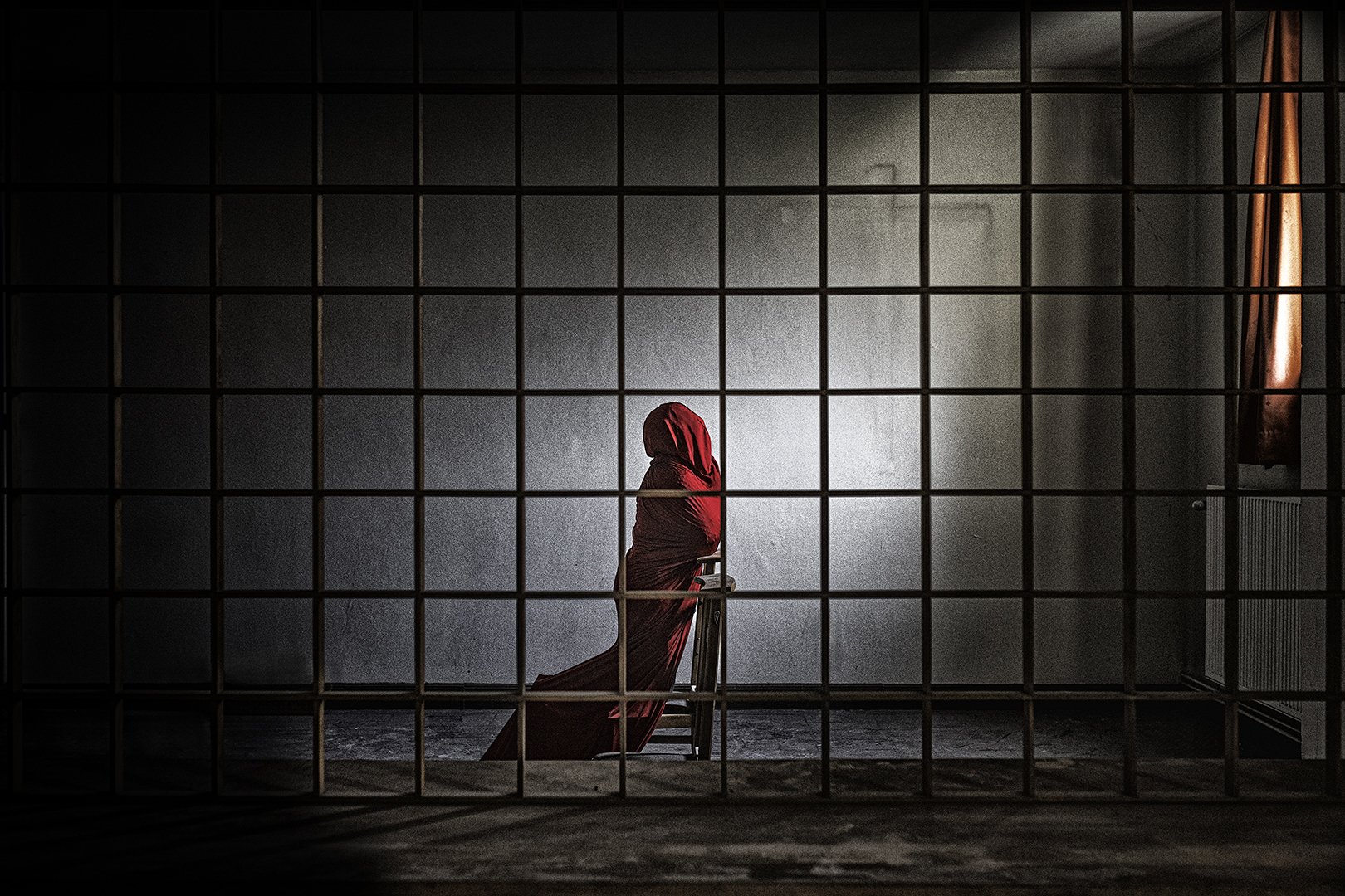 Une femme en robe rouge avec un voile couvre sa tête, vue de profil, assise sur une chaise derrière une grille en métal dans une pièce vide, avec un mur gris en arrière-plan et une fenêtre avec un rideau rouge à droite.