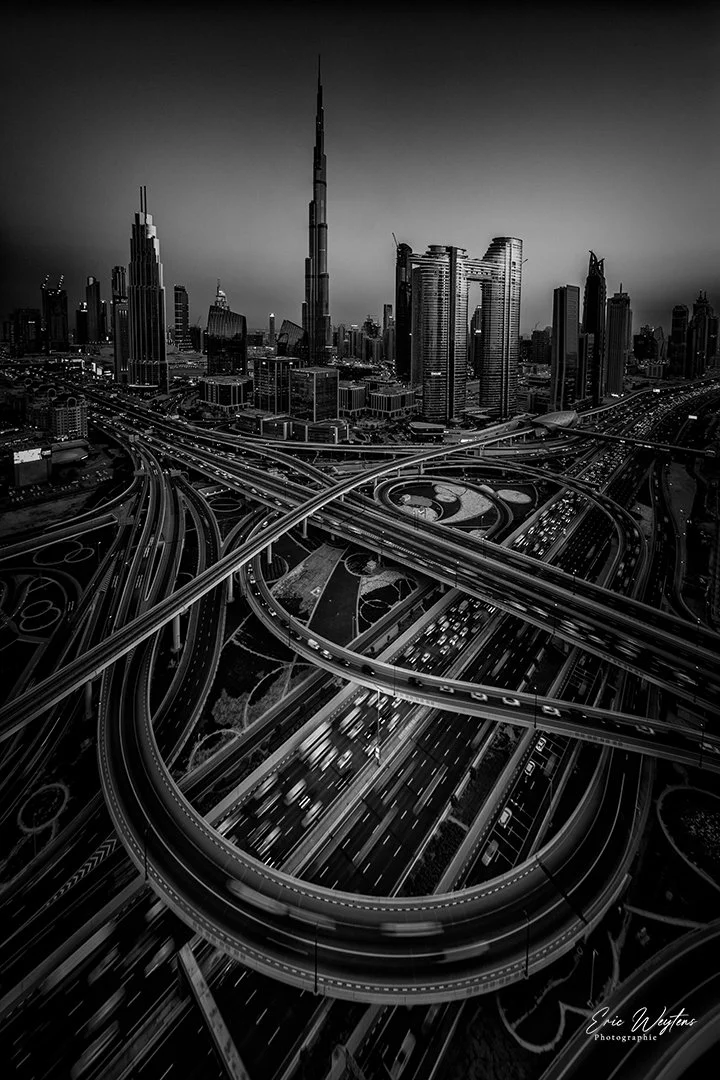 Une vue aérienne en noir et blanc d'une ville moderne avec de nombreux gratte-ciel, notamment la tour Burj Khalifa, et un réseau complexe d'autoroutes enchevêtrées en contrebas.