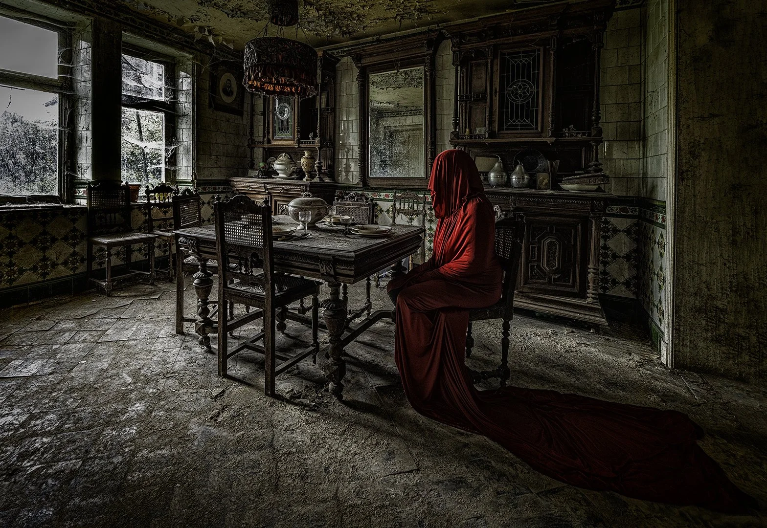 Une pièce ancienne en désarroi avec un mobilier en bois sombre, une table dressée, et une femme assise vêtue d'une robe rouge avec un voile couvrant sa tête, dont la longue traîne repose par terre. La pièce est éclairée par la lumière naturelle venan
