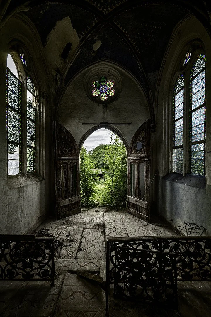 Intérieur d'une ancienne chapelle en ruines avec vitraux colorés et porte ouverte sur un jardin verdoyant extérieur.