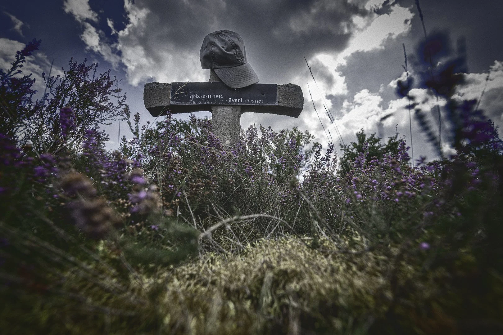 Un monument funéraire en pierre avec une casquette posée dessus, entouré de fleurs sauvages sous un ciel nuageux.