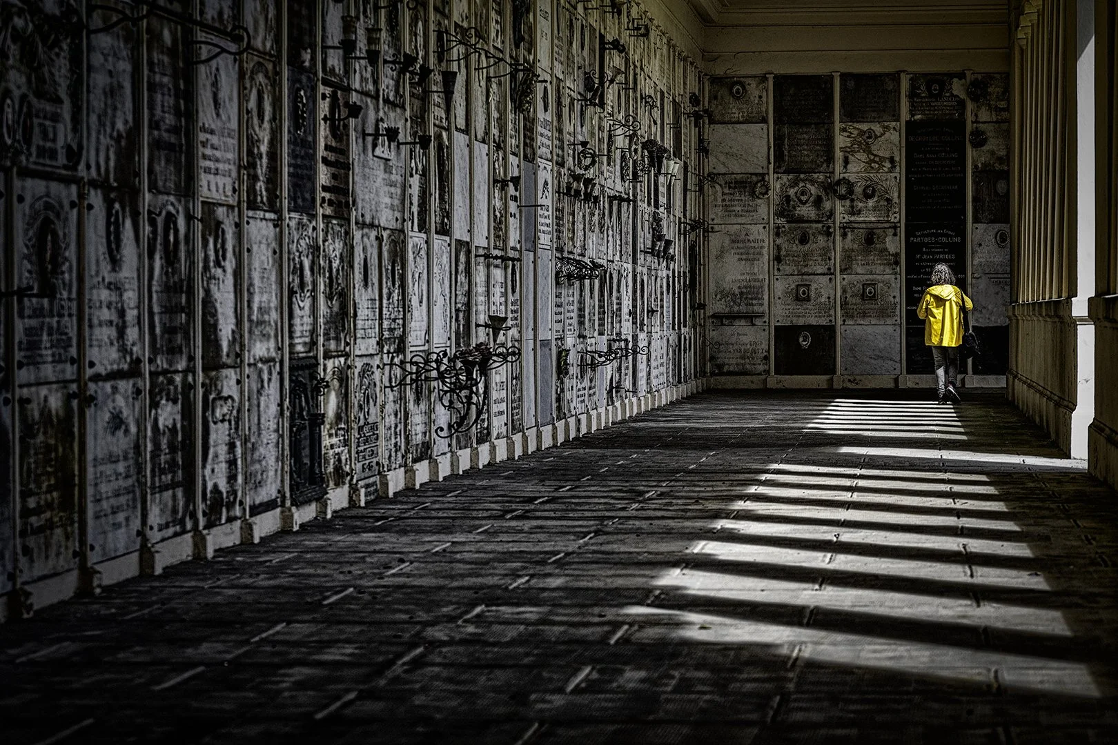 Une personne portant un manteau jaune marche dans un couloir sombre bordé de murs avec des plaques funéraires en marbre et des croix en métal.