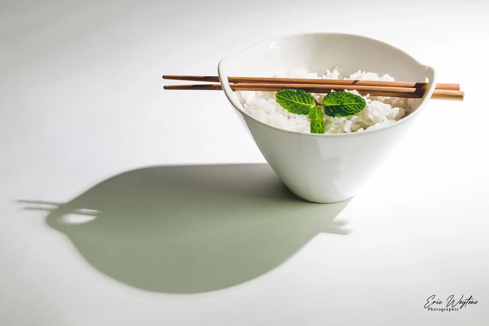 Bol de riz blanc avec des feuilles de menthe et des baguettes en bois posé dessus, projetant une ombre sur fond blanc.