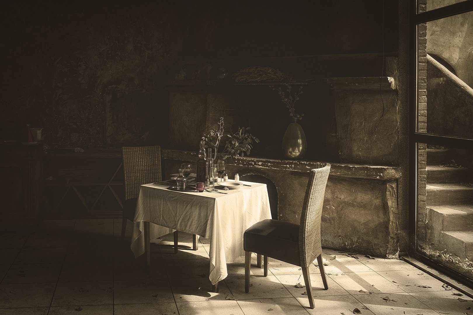 Table dressée dans une pièce sombre avec une cheminée en arrière-plan, deux chaises en rotin, une avec un siège rembourré, et une table avec une nappe blanche, de la vaisselle, des verres, des bouteilles et des vases modulés