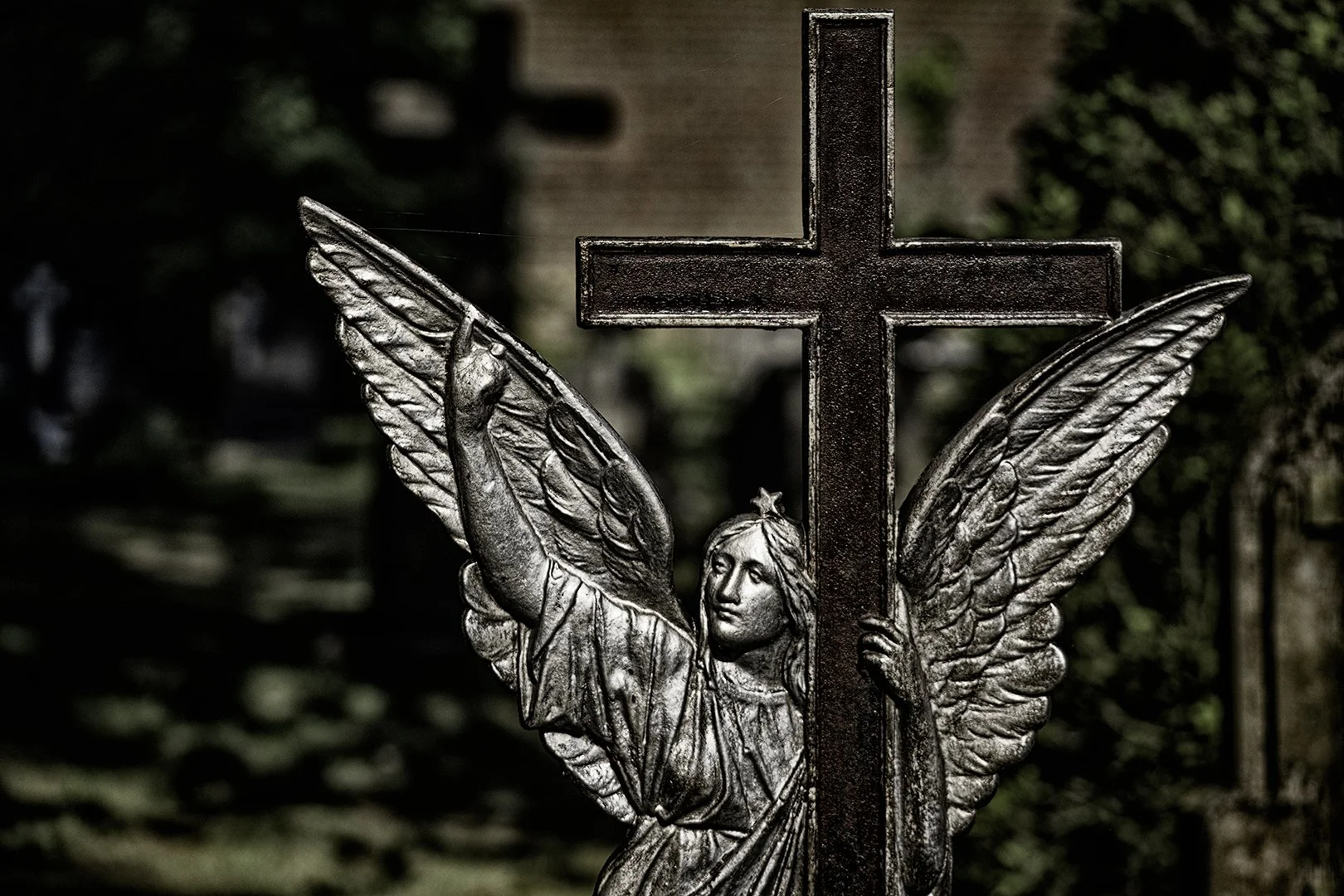 Sculpture en métal représentant un ange avec des ailes déployées, tenant une croix sur un fond flou de végétation.