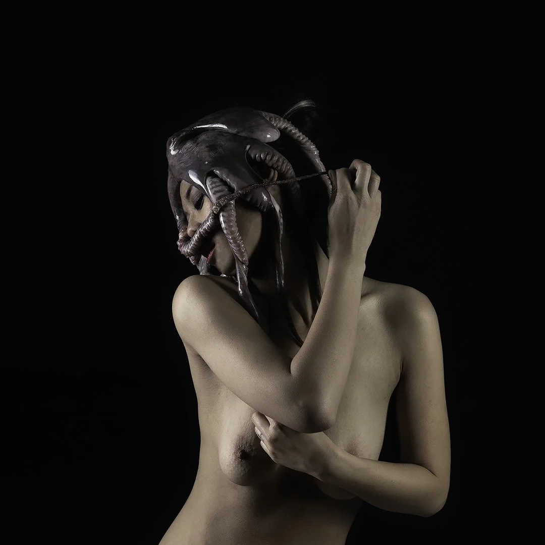 Une femme nue porte un masque ressemblant à une grande pieuvre ou un calmar, couvrant son visage et la moitié de sa tête, dans un fond noir.