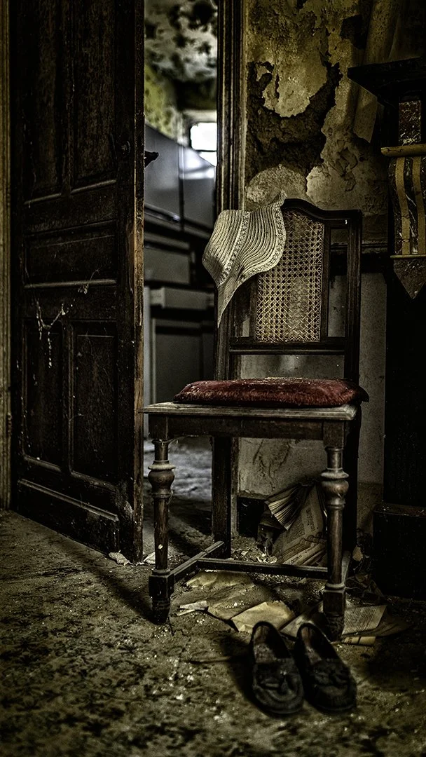 Chaise ancienne en bois avec coussin rouge, vêtements usés accrochés dessus, chaussures noires en dessous, dans une pièce abandonnée avec murs endommagés.