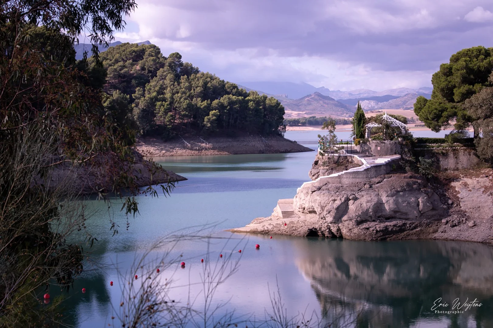 Paysage avec lac, rochers, arbres verts et une petite structure blanche sur un rocher, montagnes en arrière-plan, ciel nuageux.