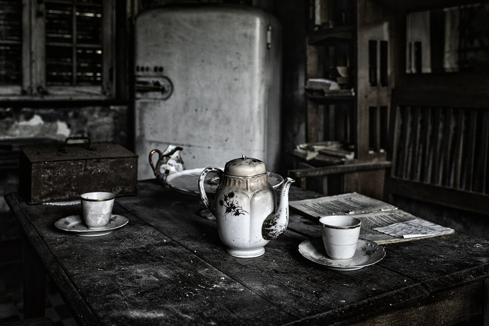 Vaisselle ancienne sur une table en bois usée dans une pièce sombre, y compris une théière blanche, deux tasses, une assiette, un vieux livre, et une boîte en métal rouillée.