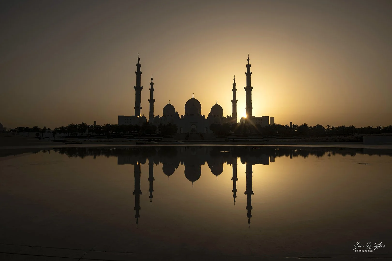 Une silhouette de mosquée avec plusieurs minarets, reflet dans l'eau au coucher du soleil.