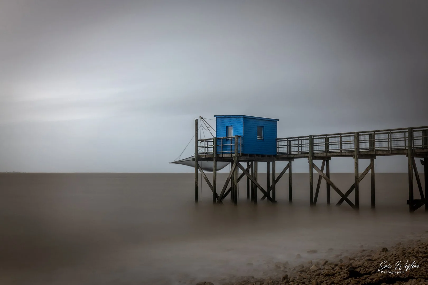 Une cabane bleue sur une jetée en bois sur la mer, avec un ciel nuageux.